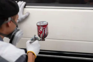 Técnico aplicando pintura automotriz de color rojo con pistola HVLP sobre la puerta de un auto, usando equipo de protección y guantes