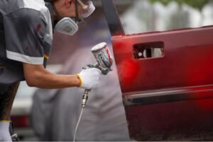 Técnico aplicando pintura automotriz de color rojo con pistola HVLP sobre la puerta de un auto, usando equipo de protección y guantes